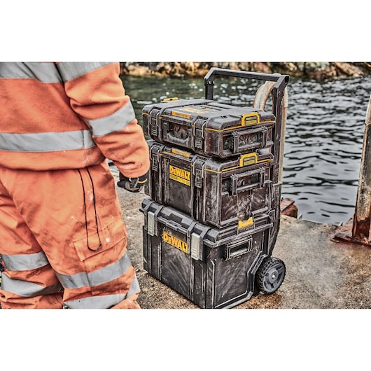 A person wearing an orange and reflective work outfit stands beside a stack of three black and yellow DEWALT tool storage
