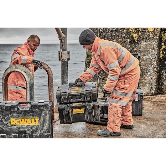 Two workers in orange safety gear are handling DEWALT ToughSystem tool storage cases on a dock by the sea. Multiple black