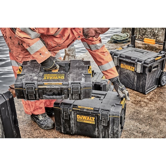 A worker in an orange safety suit and gloves is handling DEWALT ToughSystem 2.0 tool storage boxes on a construction site