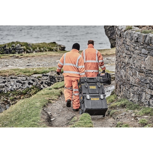 Two workers wearing orange high-visibility jackets are walking outdoors near a stone wall and carrying stacked DEWALT