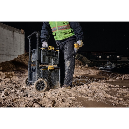 A person standing on a muddy construction site at night, wearing a green safety vest and gloves, next to a stacked DEWALT