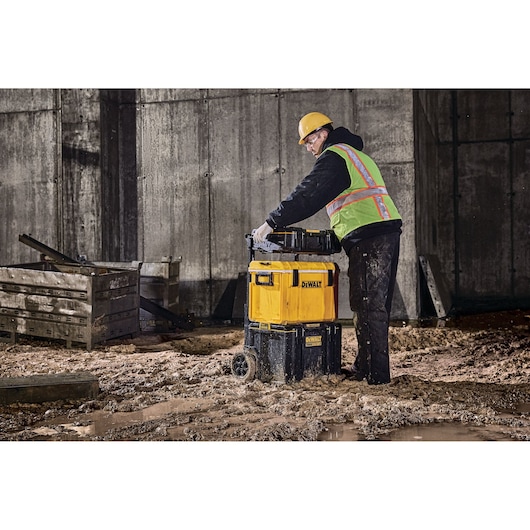 A person wearing a hard hat and high-visibility vest is working in a muddy construction site, handling a stackable yellow