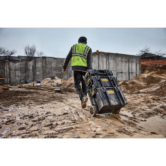 A person wearing a safety vest and helmet is pulling a DEWALT mobile tool storage box with wheels across a muddy
