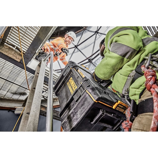 A person in a green jacket is holding a DEWALT DWST83294-1 tool storage box while ascending a metal ladder in an industrial