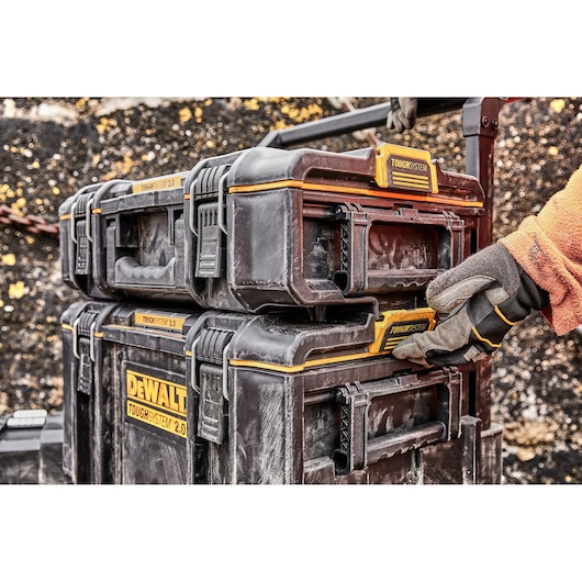 A gloved hand locks stacked DEWALT ToughSystem 2.0 toolboxes. Boxes are black with yellow branding.