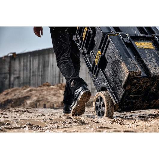 A person is pulling a muddy DEWALT ToughSystem 2.0 wheeled tool box through a wet and muddy construction site.