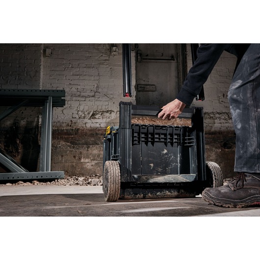 A person is pulling a DEWALT wheeled tool box (DWST83295-1) by its handle in a workshop, with visible dust and dirt on the