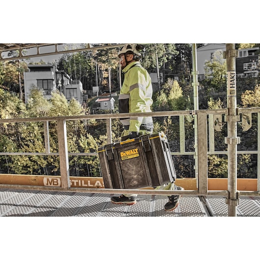 A person wearing safety gear and a helmet is carrying a DEWALT ToughSystem 2.0 storage box on a metal walkway at a construction site. Trees and buildings are visible in the background.