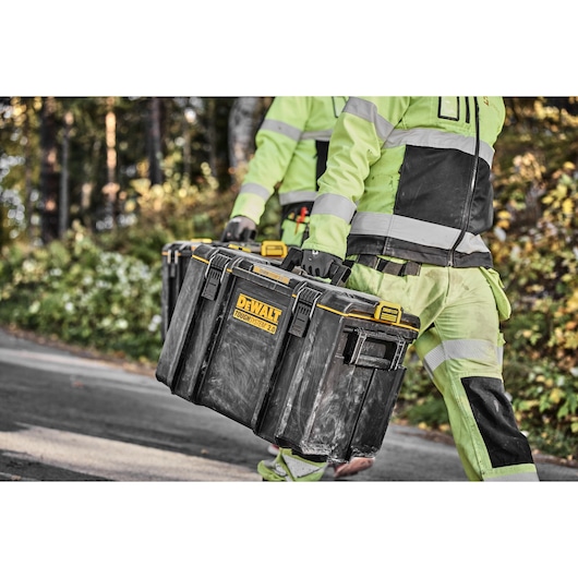 Two people in high-visibility workwear are carrying a DEWALT ToughSystem 2.0 tool storage box outdoors, with a forested background.
