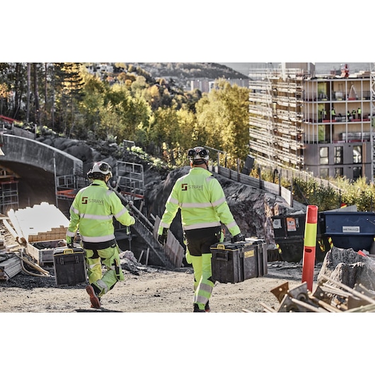 Two construction workers in high-visibility clothing and helmets are carrying DEWALT toolboxes at a construction site. There are building materials, scaffolding, and partially constructed buildings in the background.