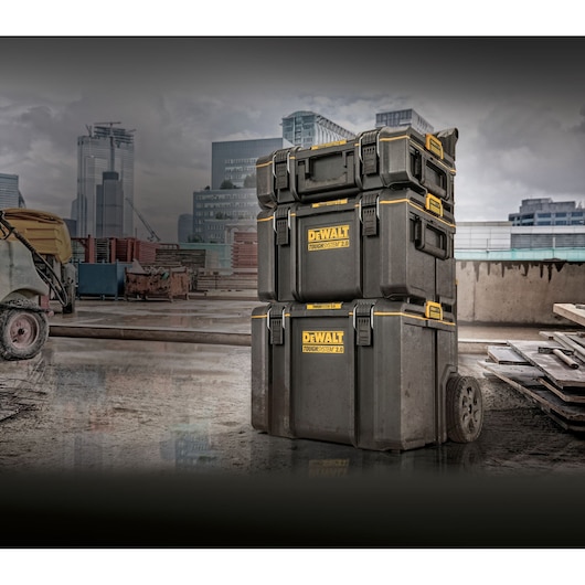 Three stacked DEWALT TOUGHSYSTEM 2.0 toolboxes with yellow accents are positioned on a construction site, with modern city buildings in the background. The bottom toolbox has wheels for portability.