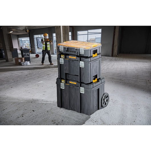Stacked DEWALT toolboxes with wheels on a construction site floor, worker in background wearing safety gear.