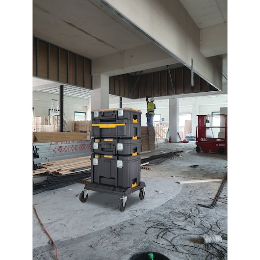 Stacked DEWALT toolboxes on wheels in a construction site with materials and worker in the background.