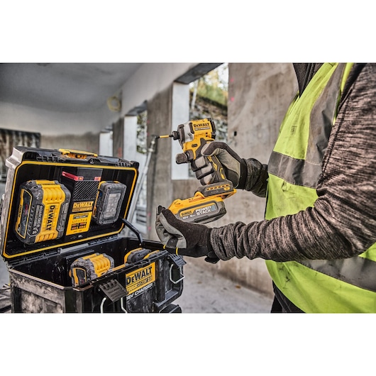 A person wearing gloves and a high-visibility vest using a DEWALT cordless power tool, with a DEWALT ToughSystem tool storage box and several battery packs visible in a construction site setting.