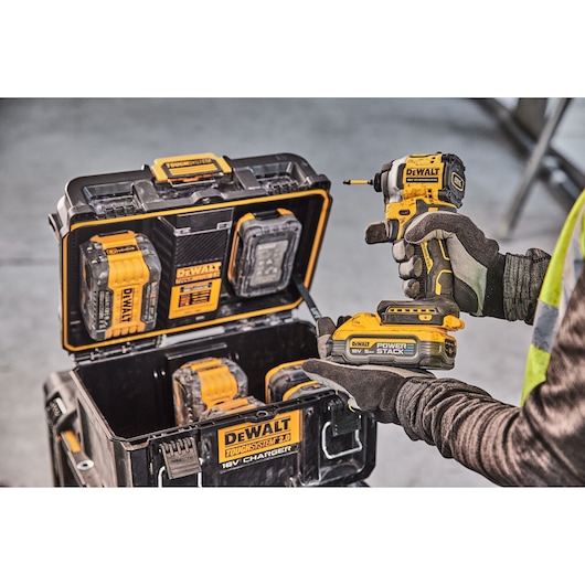 A person wearing gloves holds a DEWALT cordless power tool with a POWERSTACK battery, next to an open DEWALT ToughSystem 2.0 case containing batteries and chargers.