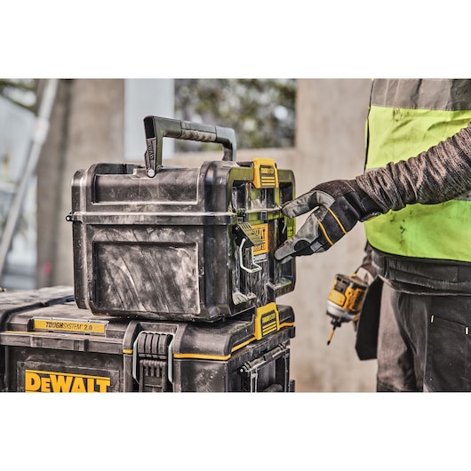 A person wearing work gloves is opening a DEWALT ToughSystem 2.0 tool storage box, which is stacked on another DEWALT box at a construction site.