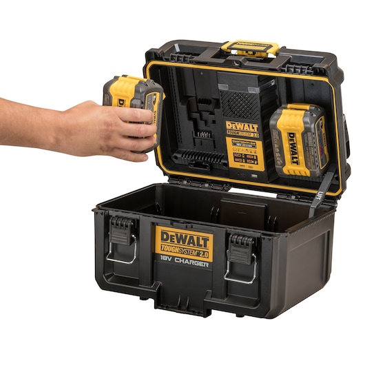 A DEWALT ToughSystem 2.0 18V Charger storage box with two batteries mounted inside the lid, and a person's hand placing a battery inside the charger compartment. The box is open showing its interior.