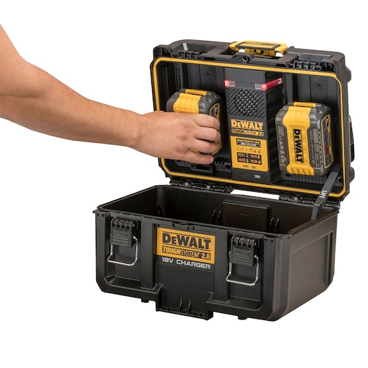A DEWALT ToughSystem 2.0 18V charger case with the lid open, showing two rechargeable batteries being placed or removed, with a hand interacting with one of the batteries. The case is black with yellow accents and labeled with DEWALT branding.