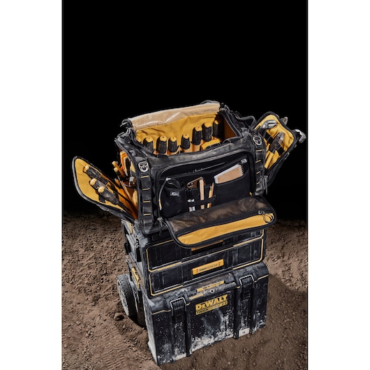 DEWALT tool bag with compartments, open and filled with tools, sitting on stacked DEWALT cases on dirt.
