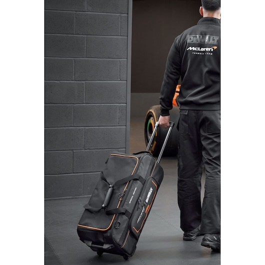 Person pulling a DEWALT McLaren wheeled tool bag in a garage with a race car visible in the background.