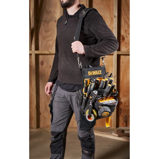 Person carrying a DEWALT tool bag with various hand tools inside, standing in front of a wooden wall.