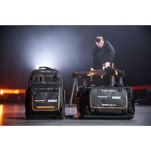 Two DEWALT McLaren tool bags on floor, one backpack and one duffel, with person working at table in background.