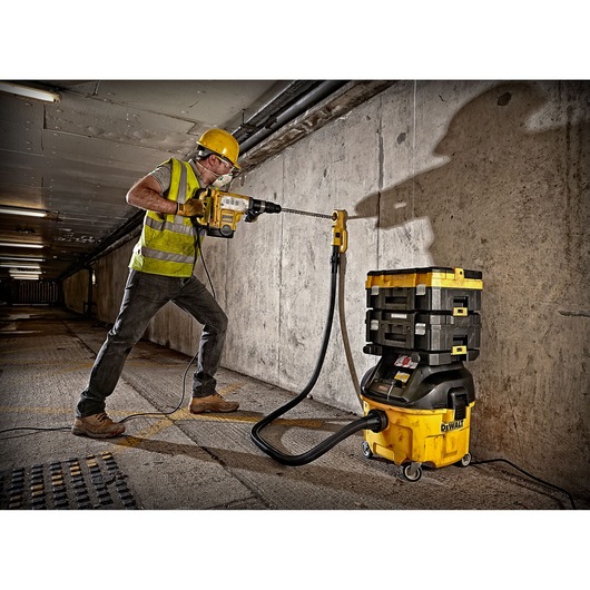 A construction worker wearing a yellow hard hat and reflective vest uses a DEWALT power tool to drill into a concrete wall in an industrial setting. A yellow DEWALT dust extractor with stacked toolboxes is connected to the drill via a hose.