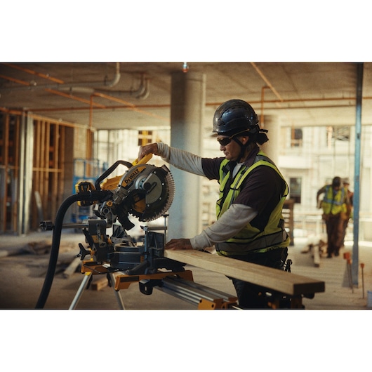 A construction worker wearing safety gear operates a DEWALT miter saw (model DCS782N) to cut a piece of wood on a jobsite. The site is indoors and still under construction, with scattered building materials and exposed structure visible in the background.
