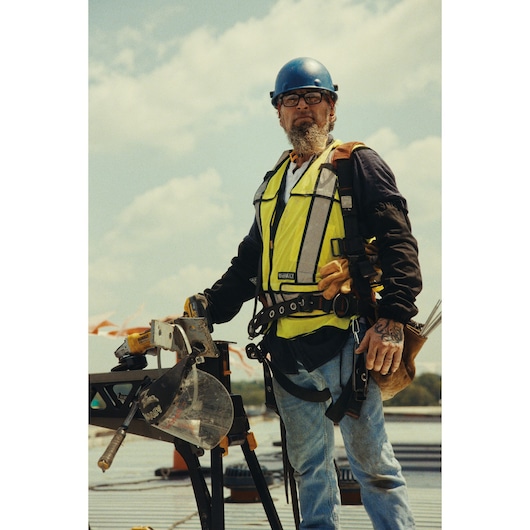 A construction worker wearing a blue safety helmet, high-visibility vest, and safety harness is standing on a worksite outdoors. The person is holding a DEWALT cordless miter saw (model DCS782N) set on a workbench. Construction materials and equipment are visible in the background.