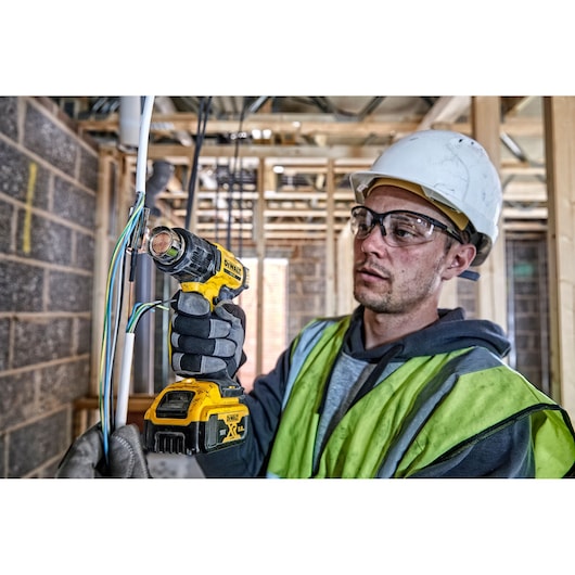 A construction worker wearing a safety helmet, gloves, and a high-visibility vest uses a yellow cordless heat gun to work on electrical wires inside a building under construction.