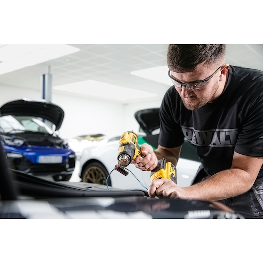 A person using a yellow heat gun while working on a car in a garage. Several cars with open hoods are visible in the background.