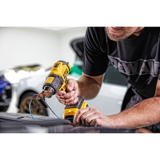 A person using a yellow cordless heat gun, branded DEWALT, to apply heat to wires or materials on a work surface. The scene appears to be in a garage with cars visible in the background.