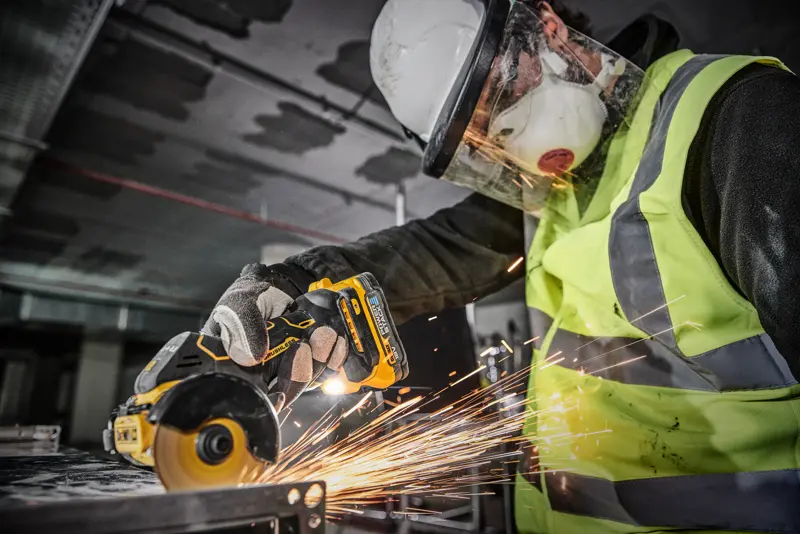 En person iført sikkerhedsudstyr, inklusive hjelm, ansigtsskærm, maske, handsker og refleksvest, bruger et DEWALT DCS438N batteridrevet værktøj til at skære metal, og det skaber gnister.