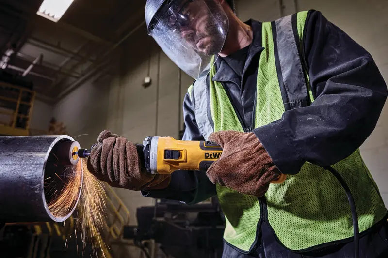 Worker using a DEWALT grinder on a metal pipe, wearing gloves, safety vest, and face shield with sparks flying.