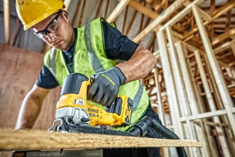 Photo of DEWALT DCS334P2 cordless jigsaw in use cutting wood at construction site, worker wearing safety gear.